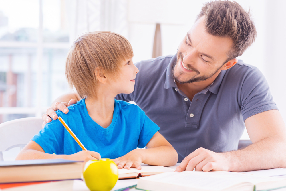 cheerful young father helping his son with homework while sitting at the table together